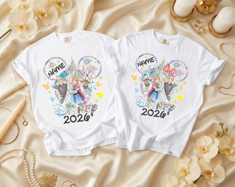 May include: Two white t-shirts featuring a colorful Disney design. The design includes the text "NAME" above a castle and characters from the movie Frozen, with the year "2026" below. One shirt has a red bow.