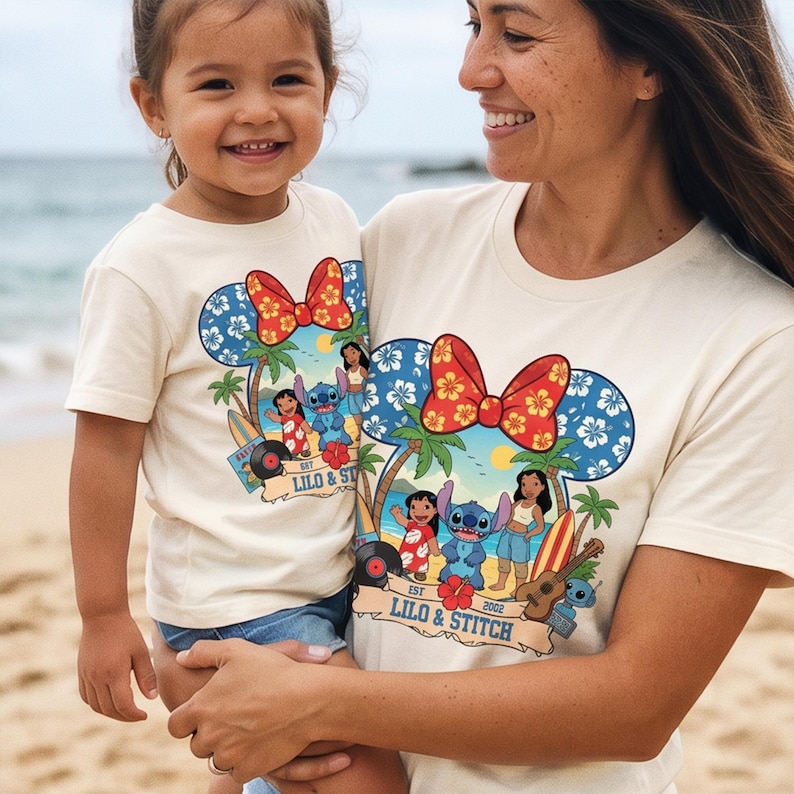 May include: Off-white t-shirts featuring a colorful graphic of Lilo & Stitch characters, palm trees, and a surfboard. The design includes a Mickey Mouse ear shape with a red bow and blue floral pattern. The text "Lilo & Stitch" is also visible.