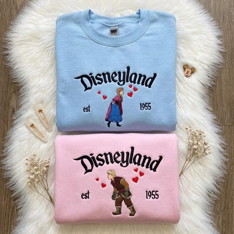 Puede incluir: Dos sudaderas, una azul claro y otra rosa, con "Disneyland" bordado en escritura negra. Cada una presenta un personaje de la pel&iacute;cula Frozen, el a&ntilde;o "1955" y la palabra "est". Las sudaderas est&aacute;n sobre una superficie blanca y esponjosa.