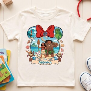 May include: White short-sleeved t-shirt featuring a colorful graphic of characters from the Disney movie Moana. The design includes Moana, Maui, Pua, Hei Hei, and other elements. The shirt has a round neckline and is made of a soft fabric.