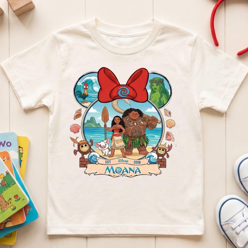 May include: White short-sleeved t-shirt featuring a colorful graphic of characters from the Disney movie Moana. The design includes Moana, Maui, Pua, Hei Hei, and other elements. The shirt has a round neckline and is made of a soft fabric.