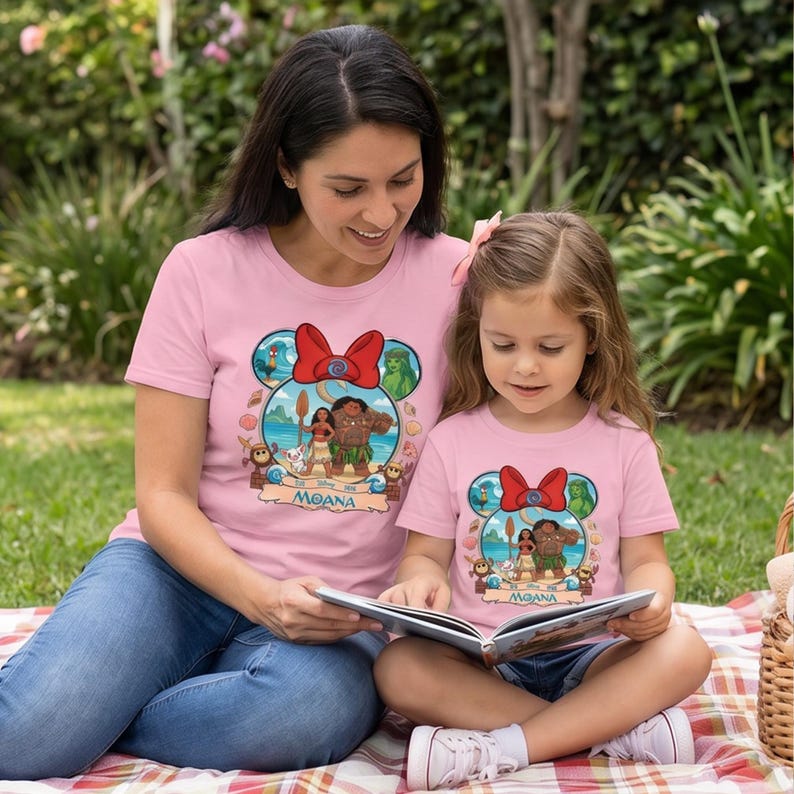 May include: Pink t-shirts featuring a Moana graphic, with a red bow and green accents. The shirts are worn by a woman and a child, who are reading a book together. The Moana graphic includes characters from the movie.
