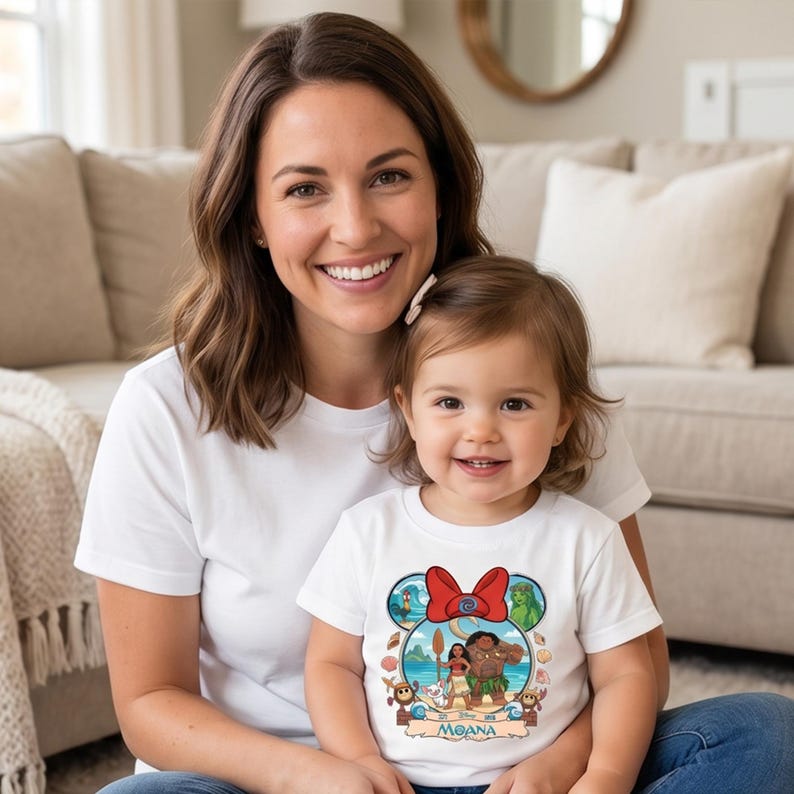 May include: A white t-shirt featuring a colorful graphic of Moana characters, including Moana, Maui, and Pua. The design is set within a Mickey Mouse-shaped frame with a red bow. The shirt is worn by a child, and the image shows a mother and child smiling.