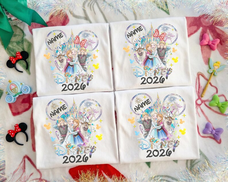 May include: Four white t-shirts with a Disney-themed graphic featuring characters, a castle, and the text "Disney 2026". Each shirt has a space for a name. Decorative bows and a fairy wand are visible.