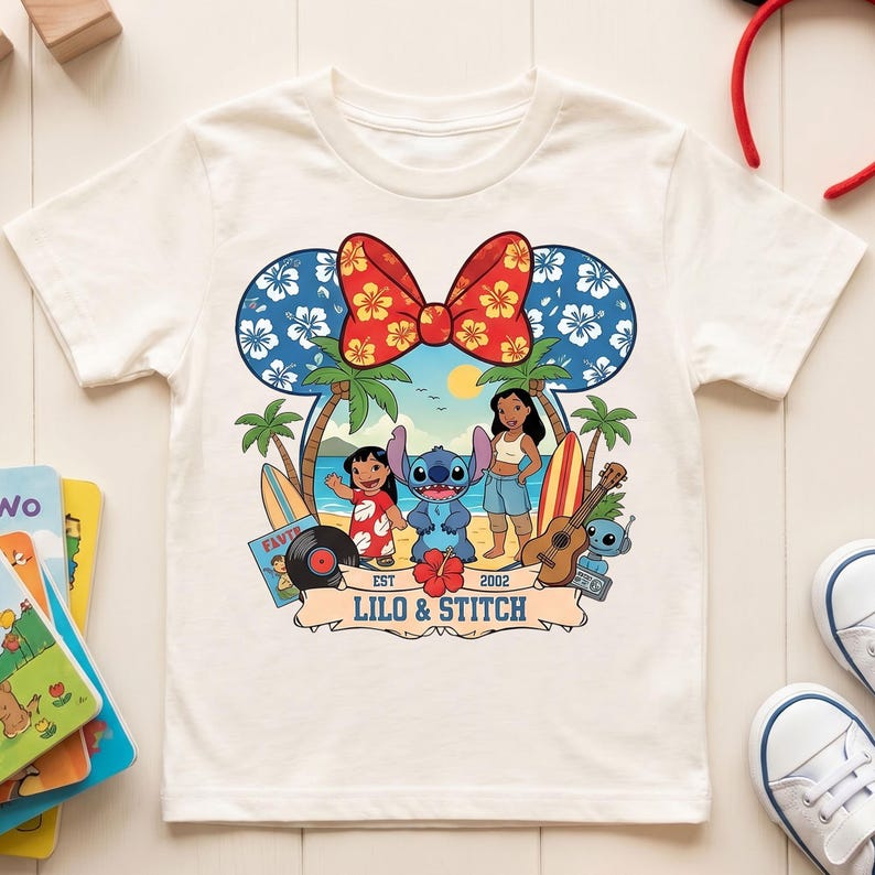 May include: White t-shirt featuring a colorful graphic of Lilo & Stitch. The design includes the characters Lilo, Stitch, and Nani, along with palm trees, surfboards, and a ukulele. The design is framed by a Mickey Mouse ear-shaped bow.