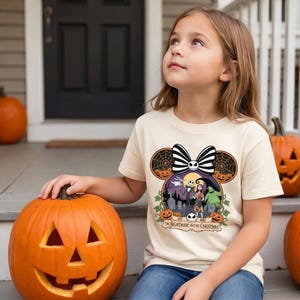 Op de afbeelding: Een cr&egrave;mekleurig T-shirt met een afbeelding van "The Nightmare Before Christmas". Het ontwerp bevat Jack Skellington, Sally en Oogie Boogie, met een Mickey Mouse oor-silhouet en een gestreepte strik. Het shirt wordt gecombineerd met een blauwe jeans.