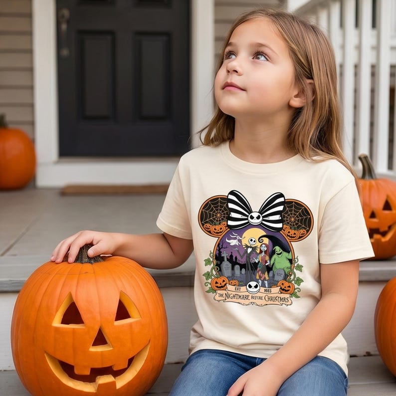 Op de afbeelding: Een cr&egrave;mekleurig T-shirt met een afbeelding van "The Nightmare Before Christmas". Het ontwerp bevat Jack Skellington, Sally en Oogie Boogie, met een Mickey Mouse oor-silhouet en een gestreepte strik. Het shirt wordt gecombineerd met een blauwe jeans.