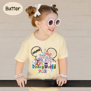 Puede incluir: Camiseta amarillo claro con un dise&ntilde;o de Disney World 2020. El dise&ntilde;o incluye un castillo, personajes y la palabra "NAME" en un dise&ntilde;o de oreja de Mickey Mouse. La camiseta la lleva un ni&ntilde;o peque&ntilde;o.