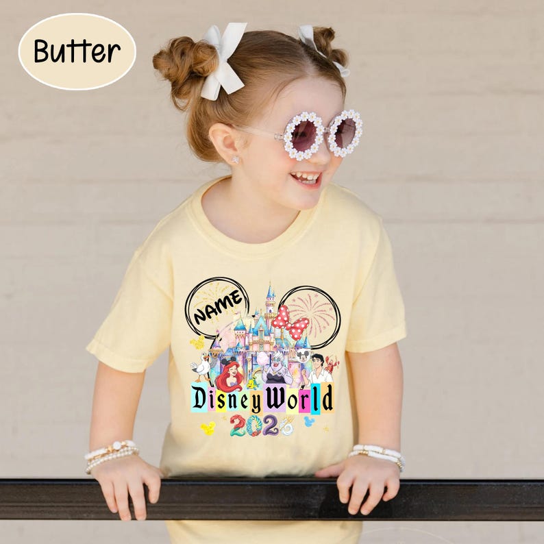 Puede incluir: Camiseta amarillo claro con un dise&ntilde;o de Disney World 2020. El dise&ntilde;o incluye un castillo, personajes y la palabra "NAME" en un dise&ntilde;o de oreja de Mickey Mouse. La camiseta la lleva un ni&ntilde;o peque&ntilde;o.