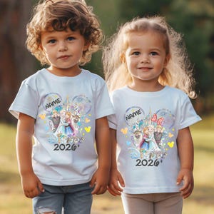 May include: Two children wearing white t-shirts with a colorful graphic design. The design features a castle, characters, and the text "NAME" and "2026". The shirts have short sleeves and are paired with blue jeans.
