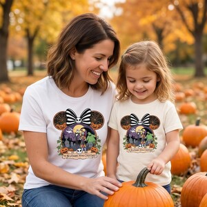 Op de afbeelding: Twee witte T-shirts met een Halloween-thema met Jack Skellington en Sally uit The Nightmare Before Christmas. Het ontwerp bevat een zwart-witte strik, spinnenwebben en pompoenen. Gedragen door een moeder en haar kind.