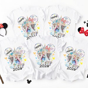 May include: White t-shirts with a colorful Disney-themed graphic featuring characters and a castle design. The shirts include the text "NAME" and "2026". Accessories include a Minnie Mouse ear headband, a bow, a wand, and Mickey Mouse silhouettes.