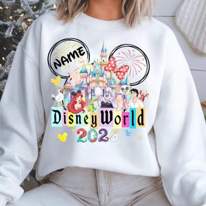 Puede incluir: Sudadera blanca con un colorido dise&ntilde;o de Disney World. El dise&ntilde;o incluye un castillo, personajes y el texto "Disney World 2023". El dise&ntilde;o tambi&eacute;n incluye dos orejas de Mickey Mouse con la palabra "NAME" en una oreja.