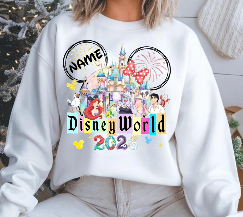 Puede incluir: Sudadera blanca con un colorido dise&ntilde;o de Disney World. El dise&ntilde;o incluye un castillo, personajes y el texto "Disney World 2023". El dise&ntilde;o tambi&eacute;n incluye dos orejas de Mickey Mouse con la palabra "NAME" en una oreja.