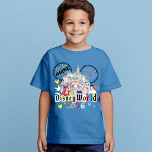 Puede incluir: Una camiseta azul con un dise&ntilde;o de Disney World. El dise&ntilde;o incluye un castillo, fuegos artificiales y varios personajes de Disney. El texto "NAME" est&aacute; encima del castillo y "Disney World 2026" debajo.