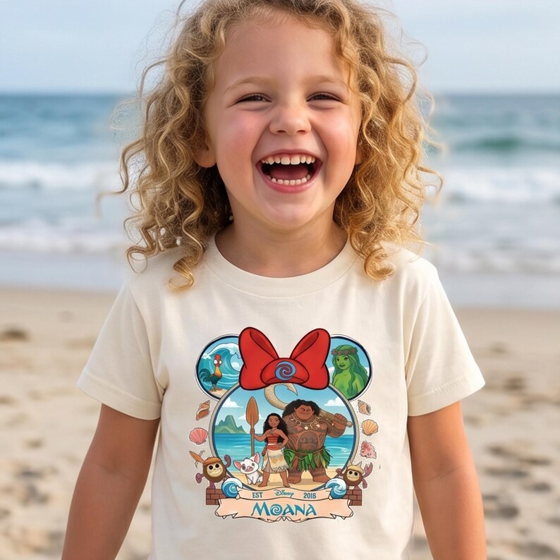 May include: A cream-colored t-shirt featuring a graphic of Moana characters within a Mickey Mouse ear design. The design includes a red bow, the word "Moana", and other characters. The child is smiling on a beach.