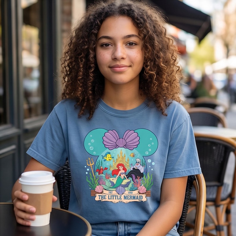 Puede incluir: Una camiseta azul con un dise&ntilde;o de La Sirenita de Disney. El dise&ntilde;o incluye a Ariel, un castillo y otros personajes dentro de una forma de oreja de Mickey Mouse. El texto "THE LITTLE MERMAID" se muestra debajo.