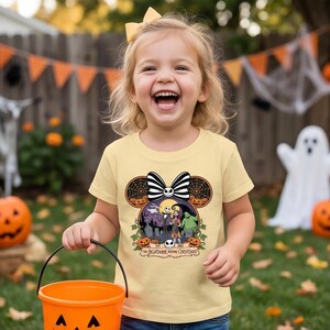 Op de afbeelding: Een kind draagt een lichtgeel T-shirt met een ontwerp van "The Nightmare Before Christmas". Het ontwerp bevat personages, pompoenen en de tekst "EST 2003". Het kind houdt een oranje Halloween-emmer vast.