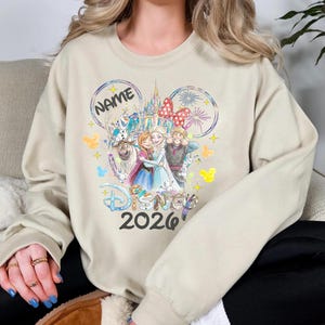 May include: Beige sweatshirt featuring a Disney-themed graphic. The design includes the word "Disney" and the year "2026", along with illustrations of characters and a castle. The word "NAME" is also present in a circular design.