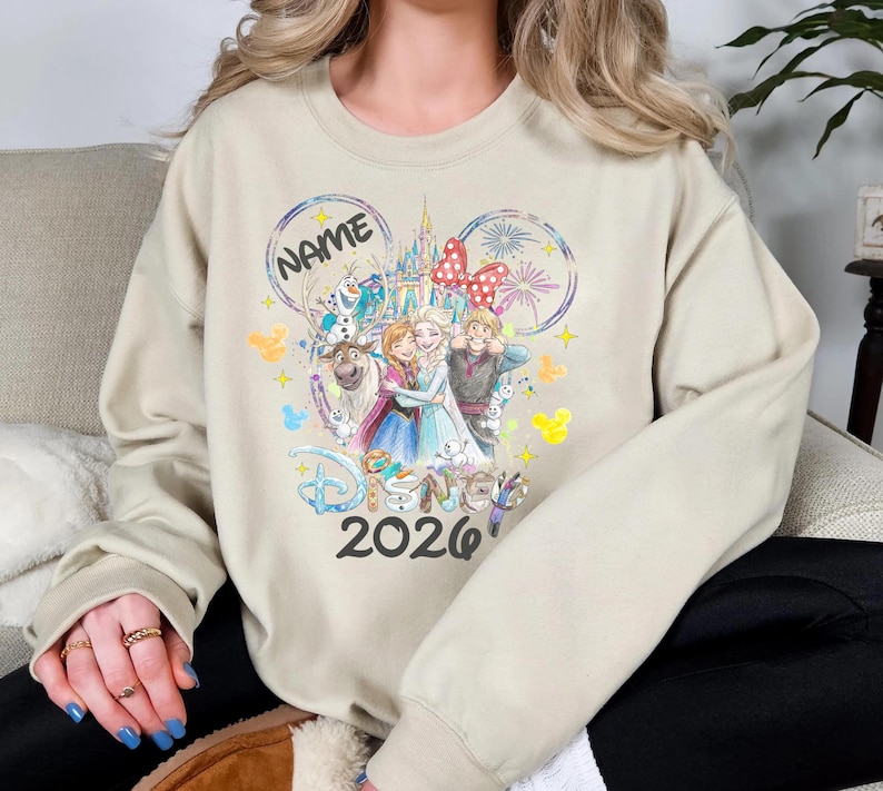 May include: Beige sweatshirt featuring a Disney-themed graphic. The design includes the word "Disney" and the year "2026", along with illustrations of characters and a castle. The word "NAME" is also present in a circular design.