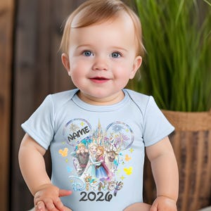May include: A light blue baby onesie featuring a Disney-themed graphic. The design includes the text "NAME" and "2026", along with images of characters and a castle. The onesie is short-sleeved.