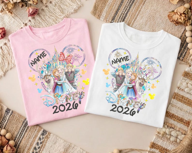 May include: Two t-shirts, one pink and one white, featuring a colorful graphic of characters from the movie "Frozen." The design includes the text "NAME" and "Disney 2026" in a whimsical font, with a castle and fireworks.