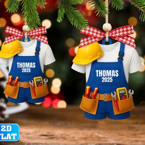 May include: Two Christmas ornaments shaped like a work uniform, featuring a white t-shirt, blue overalls, and a yellow hard hat. The overalls have the name "THOMAS" and the year "2025" printed on them. Each ornament has a red and white bow.