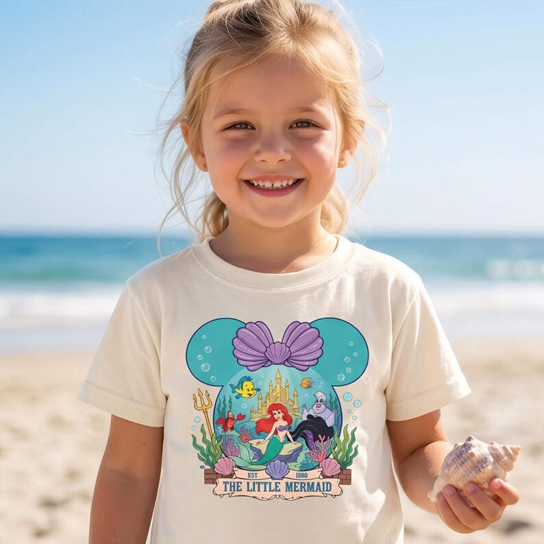 Puede incluir: Una camiseta color crema con un gr&aacute;fico de La Sirenita de Disney. El dise&ntilde;o incluye a Ariel, Flounder, Sebasti&aacute;n y &Uacute;rsula, con un castillo y el texto "EST 1989 THE LITTLE MERMAID". El dise&ntilde;o est&aacute; dentro de un contorno en forma de Minnie Mouse color turquesa.