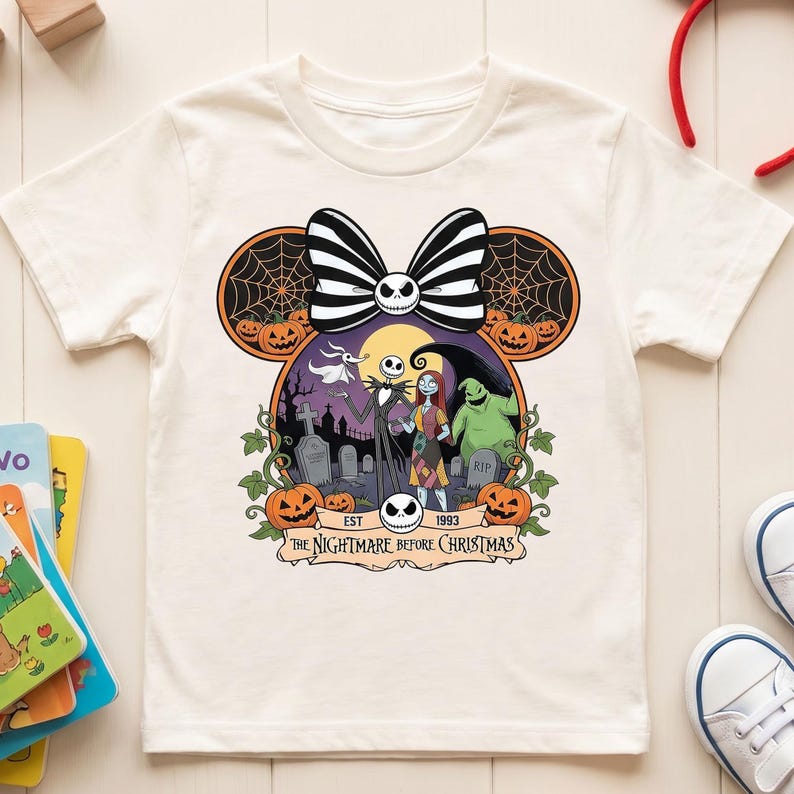 Op de afbeelding: Cr&egrave;mekleurig T-shirt met een afbeelding van "The Nightmare Before Christmas". Het ontwerp bevat Jack Skellington, Sally en andere personages, met pompoenen en een strik. De tekst "EST 1993" en "The Nightmare Before Christmas" zijn ook zichtbaar.