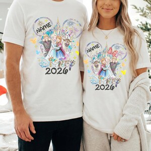 May include: Two white t-shirts featuring a colorful Disney-themed graphic. The design includes the text "NAME" in a Mickey Mouse ear shape, Disney characters, and the year "2026". The shirts are short-sleeved.