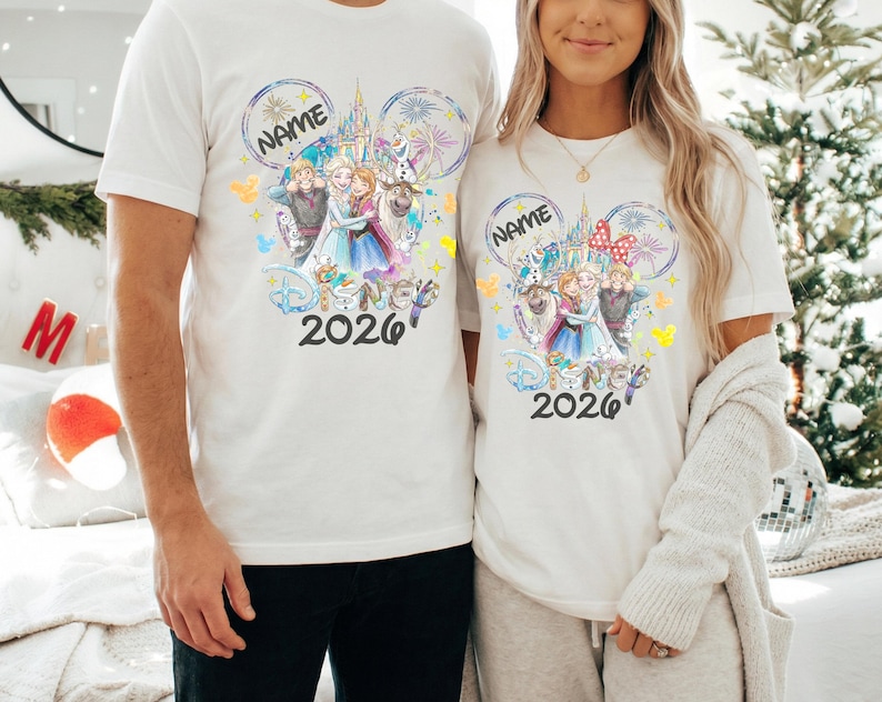 May include: Two white t-shirts featuring a colorful Disney-themed graphic. The design includes the text "NAME" in a Mickey Mouse ear shape, Disney characters, and the year "2026". The shirts are short-sleeved.