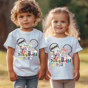 Puede incluir: Dos ni&ntilde;os con camisetas blancas con un dise&ntilde;o colorido de Disney World. El dise&ntilde;o incluye un castillo, personajes y el texto "Disney World 2026". Cada camiseta tiene un espacio para un nombre encima del dise&ntilde;o.