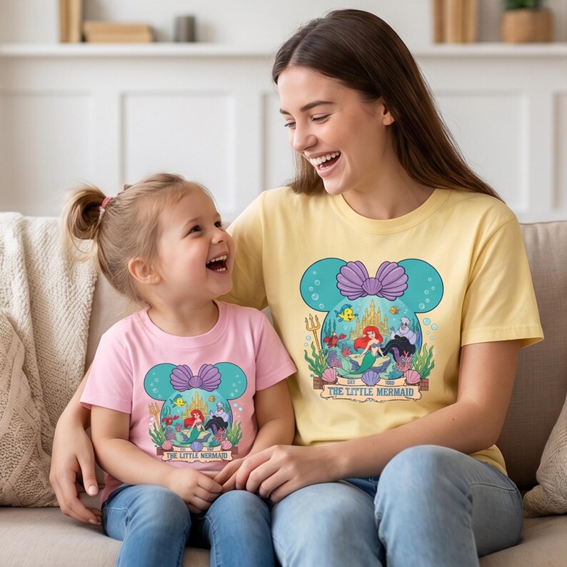 Puede incluir: Camisetas rosas y amarillas a juego con un gr&aacute;fico de Ariel de La Sirenita. El dise&ntilde;o est&aacute; dentro de una forma de cabeza de Mickey Mouse turquesa con un lazo morado. El texto "The Little Mermaid" est&aacute; debajo.