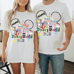 Puede incluir: Camisetas blancas con un dise&ntilde;o de Disney World 2020. El dise&ntilde;o incluye un castillo, fuegos artificiales y varios personajes de Disney. El texto "Disney World 2020" est&aacute; en letras coloridas. Las camisetas tienen un dise&ntilde;o de orejas de Mickey Mouse con la palabra "NAME" en un c&iacute;rculo.