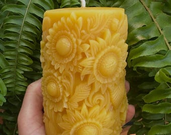 May include: A yellow beeswax candle with a sunflower design carved into the surface. The candle has a white wick and is surrounded by green fern leaves. The candle is approximately 4 inches tall.