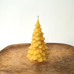 Könnte beinhalten: Eine gelbe Weihnachtsbaumkerze aus Bienenwachs. Die Kerze hat die Form eines Tannenbaums mit detaillierten, geschichteten Ästen. Sie steht auf einem Holztablett vor einem weißen Hintergrund. Die Kerze hat einen silbernen Docht.