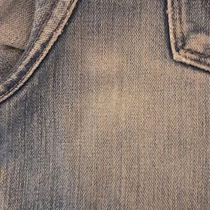 May include: Close-up of denim fabric with visible texture and stitching details. The image shows a section of jeans, including a pocket and seams, with a faded, worn appearance. The color is a light to medium blue.