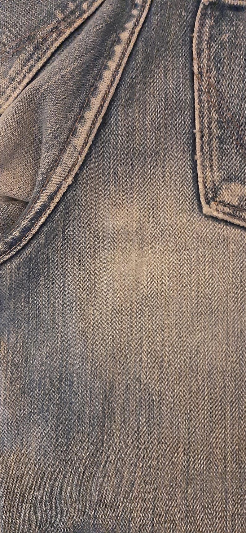 May include: Close-up of denim fabric with visible texture and stitching details. The image shows a section of jeans, including a pocket and seams, with a faded, worn appearance. The color is a light to medium blue.
