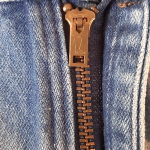 May include: A pair of light blue denim jeans featuring a copper-colored metal button at the waistband and a brass-toned zipper fly. The zipper pull is embossed with a small boot icon, and the fabric displays a classic, faded blue wash texture.