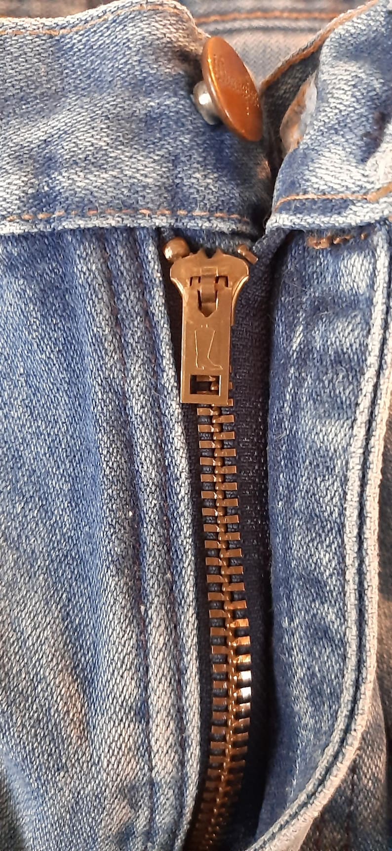 May include: A pair of light blue denim jeans featuring a copper-colored metal button at the waistband and a brass-toned zipper fly. The zipper pull is embossed with a small boot icon, and the fabric displays a classic, faded blue wash texture.