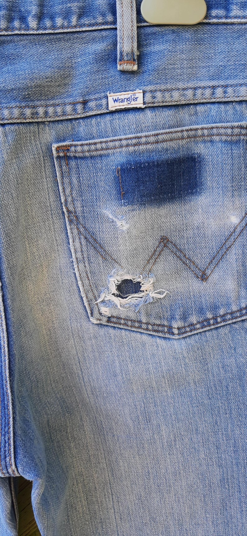 Może przedstawiać: Zbliżenie jasnoniebieskich dżins&oacute;w z zniszczoną kieszenią. Widoczna jest metka Wrangler. Dżinsy mają zużyty wygląd z naszywką i dziurą w kieszeni.