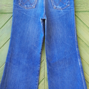 May include: Blue denim Wrangler jeans with a wide leg. The jeans have two back pockets and a classic five-pocket design. The jeans are hanging on a Prada hanger.