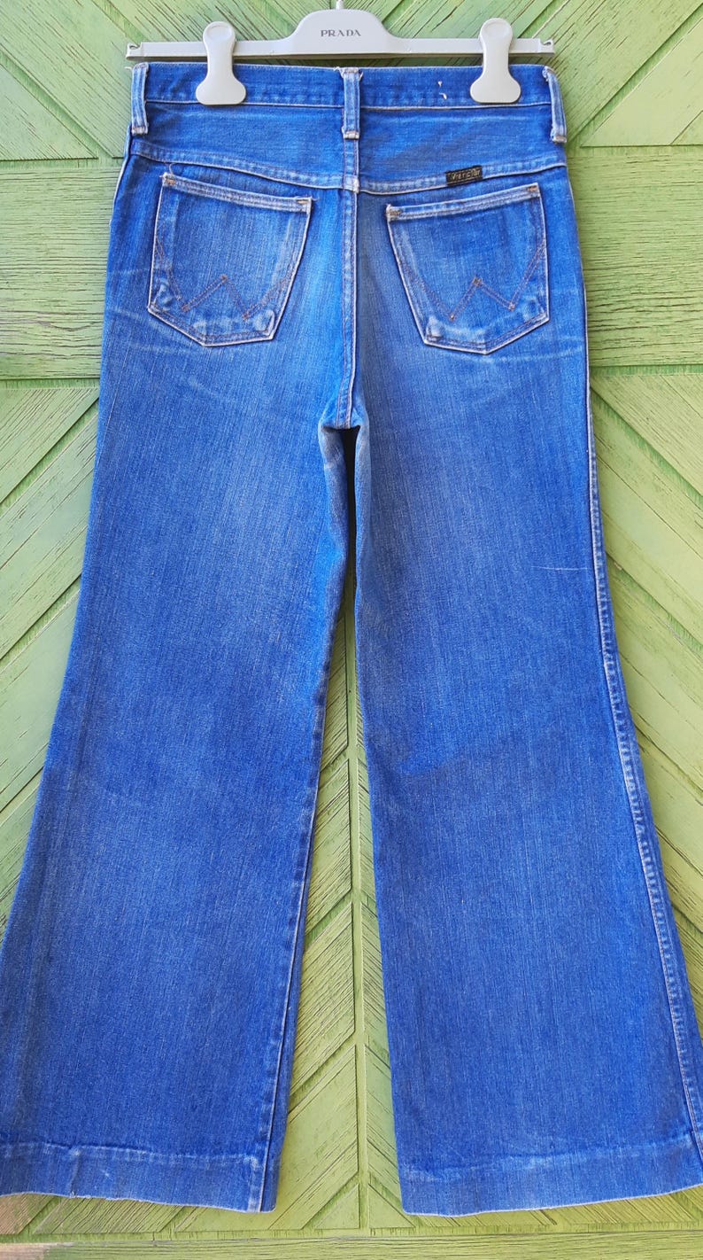 May include: Blue denim Wrangler jeans with a wide leg. The jeans have two back pockets and a classic five-pocket design. The jeans are hanging on a Prada hanger.