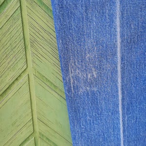 May include: A pair of blue denim jeans featuring a medium wash with subtle fading and a vertical crease running down the center of the leg. The fabric shows a textured weave with visible stitching along the hem at the bottom of the pant leg.