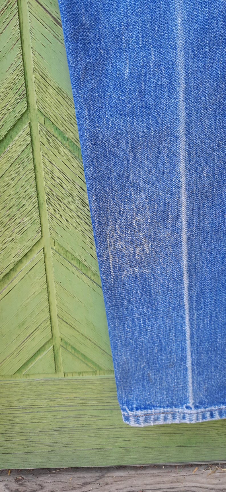 May include: A pair of blue denim jeans featuring a medium wash with subtle fading and a vertical crease running down the center of the leg. The fabric shows a textured weave with visible stitching along the hem at the bottom of the pant leg.