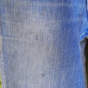 Pode incluir: Close-up de jeans de ganga azul. O tecido mostra uma lavagem desbotada com varia&ccedil;&otilde;es sutis de cor. Existem algumas pequenas manchas escuras na ganga. Os jeans s&atilde;o de um estilo cl&aacute;ssico.