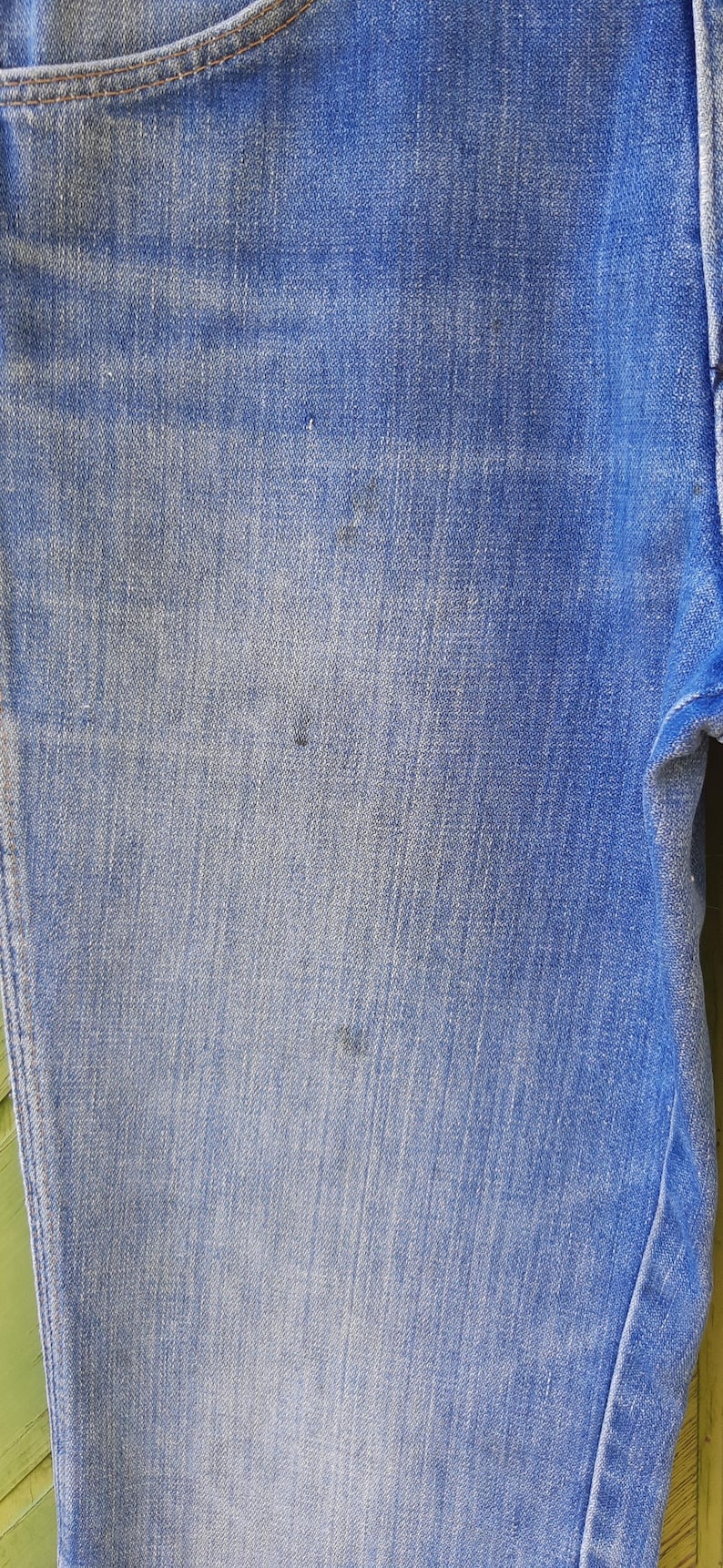 Pode incluir: Close-up de jeans de ganga azul. O tecido mostra uma lavagem desbotada com varia&ccedil;&otilde;es sutis de cor. Existem algumas pequenas manchas escuras na ganga. Os jeans s&atilde;o de um estilo cl&aacute;ssico.
