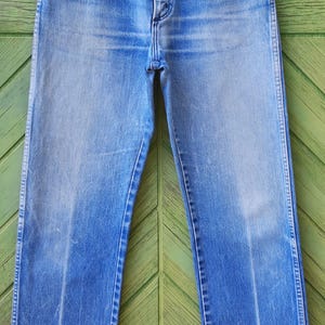 May include: High-waisted blue denim jeans featuring a classic five-pocket design, a button fly closure, and copper-toned rivets. The fabric displays a faded wash with vertical whiskering patterns along the hips and straight-cut legs.