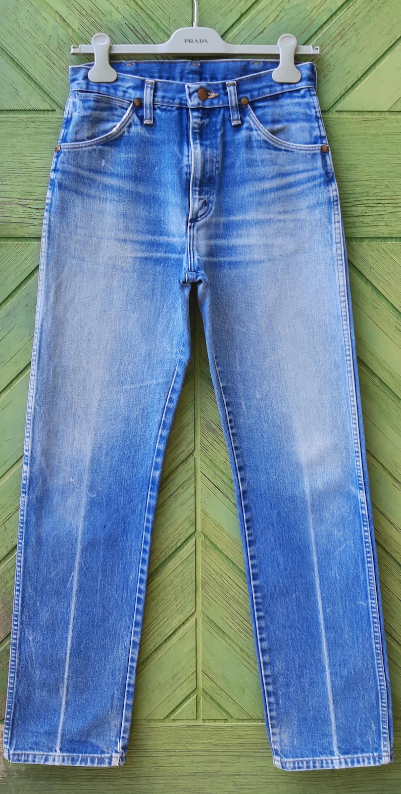 May include: High-waisted blue denim jeans featuring a classic five-pocket design, a button fly closure, and copper-toned rivets. The fabric displays a faded wash with vertical whiskering patterns along the hips and straight-cut legs.