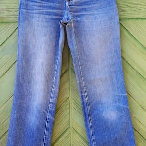 May include: A pair of light blue denim jeans with a copper button and a small pocket on the right side. The jeans are hanging on a white hanger with a "PRADA" label.
